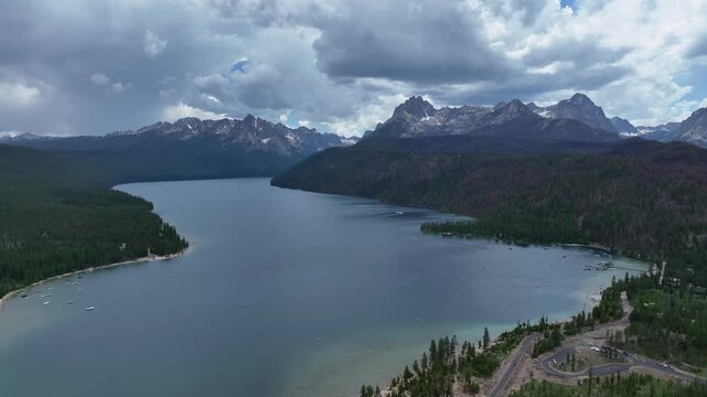 Aerial view of Redfish Lake surrounded by the Sawtooth Mountains and dense pine forests under a dramatic cloudy sky in Stanley, Idaho, United States.