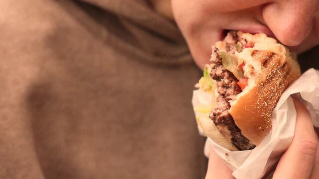 Close-up of a woman taking a big bite of juicy grilled beef burger with fresh lettuce, tomato and pickles, toasted sesame bun wrapped in white paper, blurred warm restaurant interior background scene