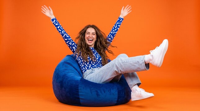 Joyful young woman wearing patterned blue shirt sits on comfortable bean bag chair with raised arms while laughing and celebrating success in this bright studio photograph.