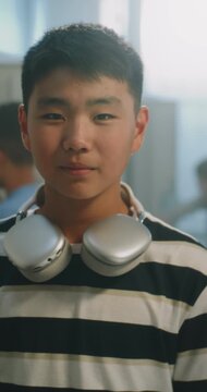 Portrait of Friendly Asian Teenage Boy Student with Wireless Headphones Waving Hand and Looking at Camera in School Corridor. Responsibility, Familiar Routines of Teenage Education. Vertical Shot.