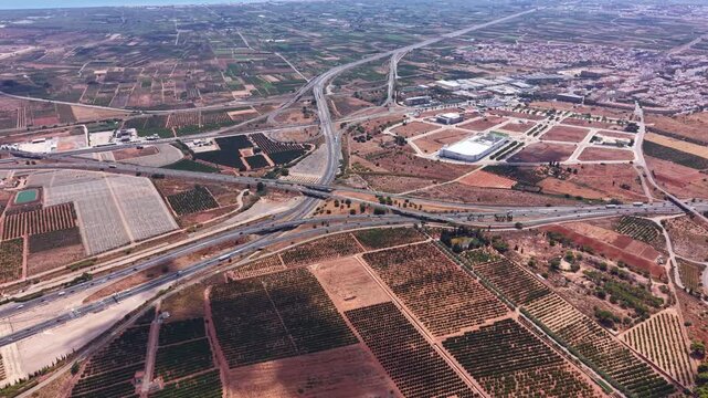 Aerial view of a major highway interchange cutting through agricultural land and orchards near a Spanish city, showing road infrastructure, farmland patterns and urban sprawl