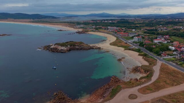 Wide aerial drone view of A Lanzada beach in Galicia Spain showing a long sandy shoreline a small rocky islet turquoise water dunes and a coastal town in the background