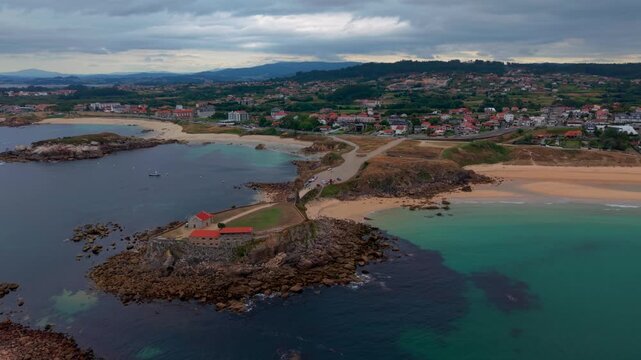 Aerial drone view over the Lanzada headland and its chapel looking toward the coastal town with beaches reefs and turquoise water in Galicia Spain