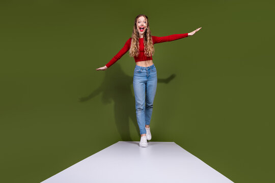 Young woman in red top and jeans balances on a white platform with green background