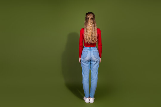 Young woman standing with red top and jeans facing away against green background