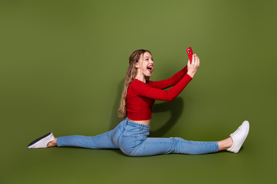 Young woman performs a full split while taking a selfie in a bright red top and jeans against a green backdrop