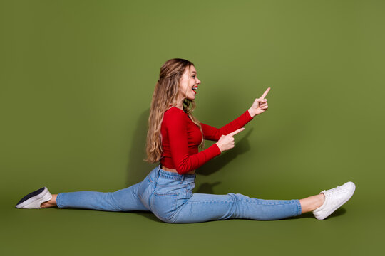 Young woman doing a full split against a green backdrop pointing and smiling in casual sportswear