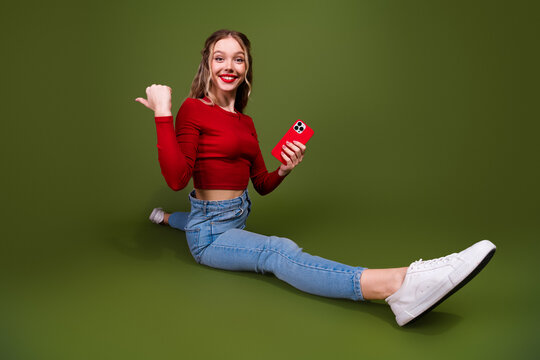 Young woman smiling sits on green studio background wearing red top jeans holding red phone for lifestyle advertising