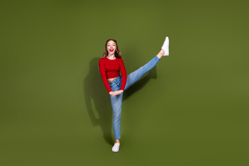 Naklejka premium Young woman in red top and jeans kicks her leg up on green studio backdrop smiling