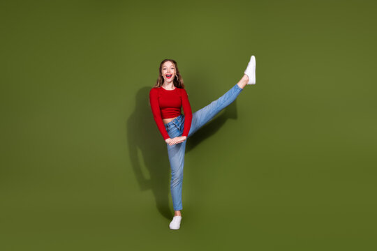 Young woman in red top and jeans kicks her leg up on green studio backdrop smiling