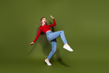 Naklejka premium Young woman jumping with joy in casual red top jeans and white sneakers against a green studio background