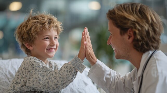 A smiling young boy in a patterned gown gives a high five to a male doctor in a white coat against a blurred background.