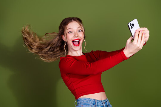 Young woman taking a selfie in a red top smiling with phone against green background