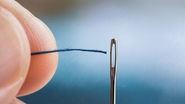 Close-up of fingers threading a blue cotton thread through the eye of a sewing needle. Macro view of hand sewing preparation and needlework hobby