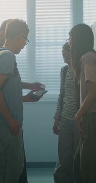 Four School Classmates Stand Together in School Locker Area, Chatting and Reacting Content on Shared Smartphone. Gen Z Lifestyle, Friendship, Digital Communication, Social Interaction. Vertical Shot.
