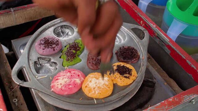 Anonymous street food vendor serving and packaging colorful kue cubit traditional Indonesian mini pancakes into a plastic container