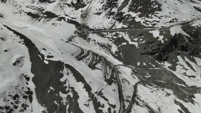Aerial view of zigzagging loop switchback roads climbing a steep rocky mountain face between snowfields at Shinku La Pass Zanskar shot by a drone flying over.
