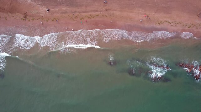 A high-angle aerial shot showing a sandy beach, incoming ocean waves, and rocks in shallow coastal water.