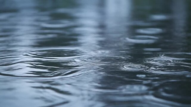 Abstract pattern of water surface disturbance caused by falling raindrops, creating ripple effects and concentric circles on a dark, reflective liquid, evoking a feeling of natural phenomenon