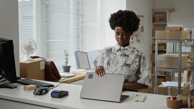 Young adult African American woman closing laptop, grabbing bag, and turning off lights in warehouse office. Clip suitable for small business, entrepreneurship, and end of workday marketing