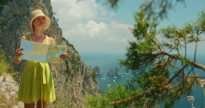 Woman in summer dress reading a map on Capri cliff with Faraglioni sea stacks behind her