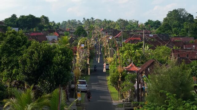 Aerial view of traditional Balinese village street decorated with penjor, featuring lush greenery, a person walking, and a scooter in Kecamatan Tegallalang, Bali, Indonesia.