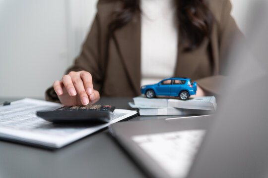 Businesswoman using a calculator beside a car model, cash and laptop, representing car insurance, auto finance, loan payments, vehicle purchase costs, budgeting, credit, and financial planning.