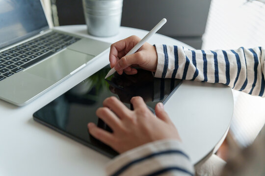 Woman using a stylus on a blank digital tablet beside a laptop, representing creative workflow, digital writing, design, productivity, remote work, planning, technology and a modern home workspace.