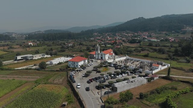 Aerial view of Igreja Paroquial de Santo Andre de Palme with its surrounding cemetery, parking lot filled with cars, and lush green fields in Palme, Braga, Portugal.