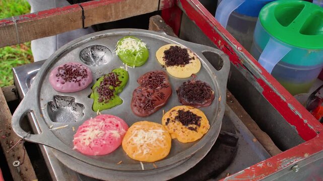 Anonymous street food vendor lifting lid revealing colorful kue cubit traditional Indonesian mini pancakes cooking on a hot iron pan