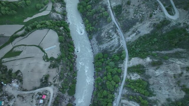 Aerial view of river flowing in mountains, past farm terraces and village houses in Jispa Lahaul Valley shot by a drone flying right above the river Bhaga in the himalayas
