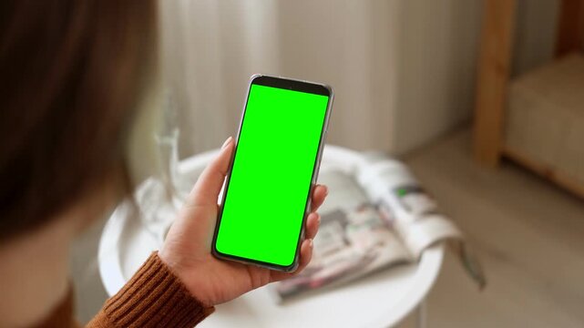 Over the shoulder view of young woman holding smartphone vertically with green screen mockup chroma key blank display. Female user looking at phone template mockup screen inside cozy room at home