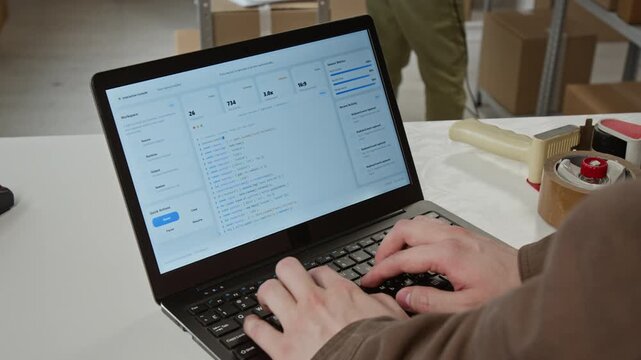 Young adult Caucasian man typing code on laptop in warehouse, managing e-commerce logistics software. High angle close up ideal for supply chain technology videos