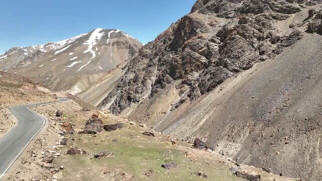 Aerial view of winding mountain road traversing dry rocky slopes below snow-streaked peaks in bright sunlight at Shinku La Pass Zanskar shot by a drone
