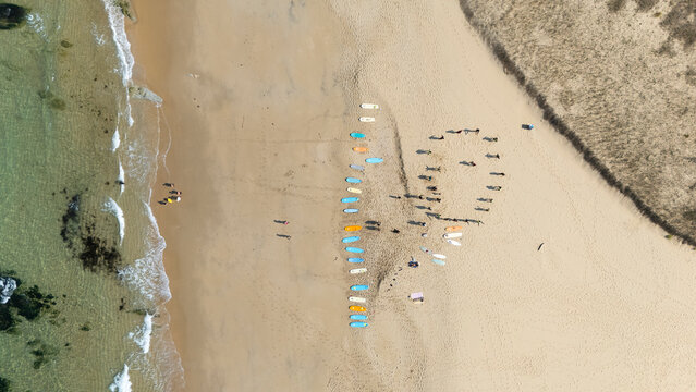 Aerial view of surf school lesson with surfboards on Atlantic beach in Spain. Colorful surfboards are lined up on the sand while a group of surfers receives instruction before entering the ocean.