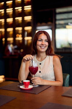 Smiling Woman Enjoying Red Cocktail in Stylish Caf&eacute; in a Casual Sleeveless Dress for Social Dining