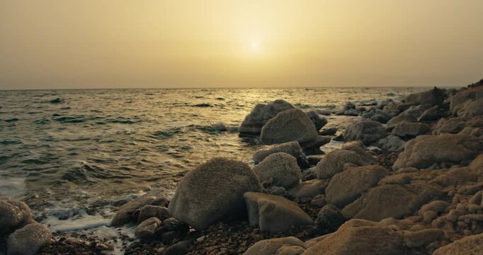 Rocky salt covered shoreline of the Dead Sea in Jordan with gentle waves, warm sunset light, and a tranquil hypersaline landscape.