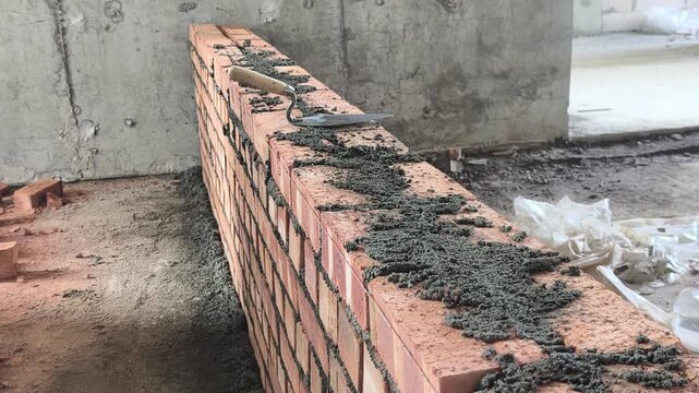 Double clay brick wall under construction on a job site with fresh wet mortar. A bricklaying trowel rests on top of the newly laid masonry line.