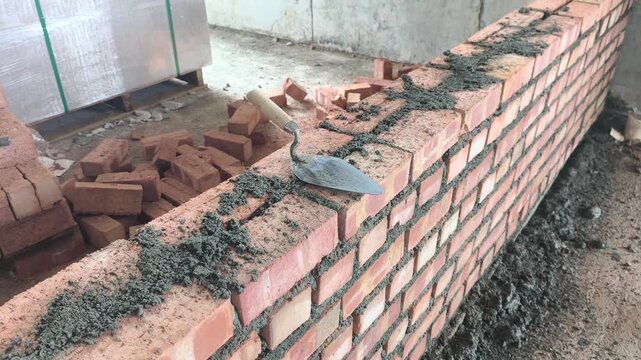 Double clay brick wall under construction on a job site with fresh wet mortar. A bricklaying trowel rests on top of the newly laid masonry line.