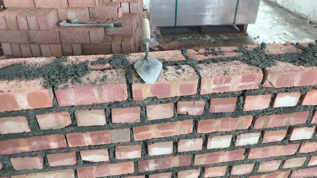 Double clay brick wall under construction on a job site with fresh wet mortar. A bricklaying trowel rests on top of the newly laid masonry line.
