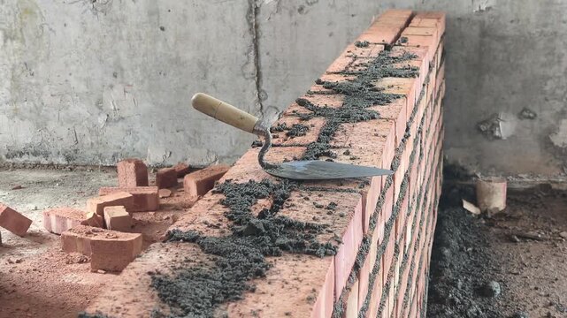 Double clay brick wall under construction on a job site with fresh wet mortar. A bricklaying trowel rests on top of the newly laid masonry line.