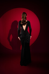 Elegant woman in a backless black evening gown standing against a dramatic red backdrop with long...