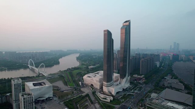 Aerial view of modern twin skyscrapers beside a winding river with a distinctive bridge in an urban cityscape.