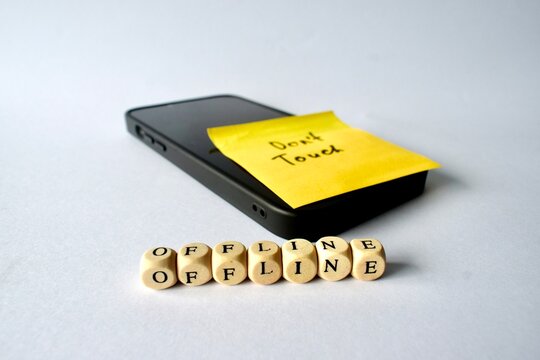 Angled shot of a smartphone with a "Don't Touch" reminder note on the screen, accompanied by wooden letter cubes spelling out "OFFLINE" on the surface.