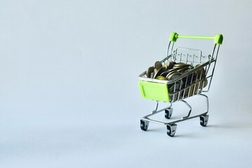 Miniature green-handled shopping cart filled to the top with mixed coins, placed on a clean background to symbolize retail spending, inflation, financial savings, and consumer purchasing power. © Akram