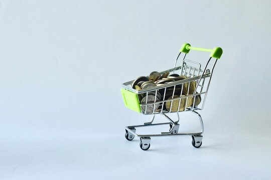 Miniature green-handled shopping cart filled to the top with mixed coins, placed on a clean background to symbolize retail spending, inflation, financial savings, and consumer purchasing power.