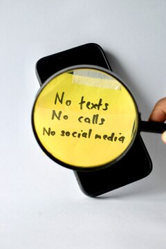 A hand holds a magnifying glass over a yellow note on a smartphone screen that reads "No texts, No calls, No social media," emphasizing a strict digital detox.