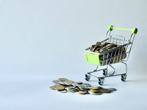 Miniature shopping cart filled with coins alongside a pile of loose change scattered on the surface, depicting retail spending, savings leakage, shopping budget, and personal finance.