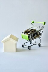 Model white house standing beside a miniature shopping cart filled with coins, symbolizing real estate costs, housing affordability, property market investment, and home savings. © Akram