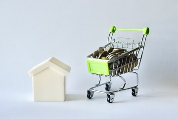 Model white house standing beside a miniature shopping cart filled with coins, symbolizing real estate costs, housing affordability, property market investment, and home savings. © Akram